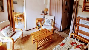 A cozy room with a wooden bed, a wooden chair, a small table, and a sofa. There is a door leading to another room with a table and chairs.