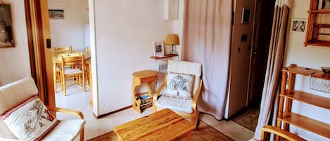 Ein gemütliches Zimmer mit einem Holzbett, einem Holzstuhl, einem kleinen Tisch und einem Sofa. Eine Tür führt in einen weiteren Raum mit Tisch und Stühlen.