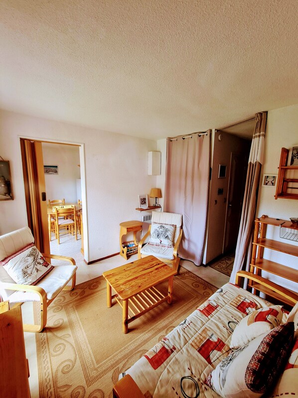 Una habitación acogedora con una cama y una silla de madera, una mesita y un sofá. Se ve una puerta que da a otra habitación con una mesa y sillas.