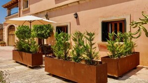 A building with large planters containing greenery, a table with chairs, and a white umbrella. - Ayurvila Wellness Retreat (Manacor)