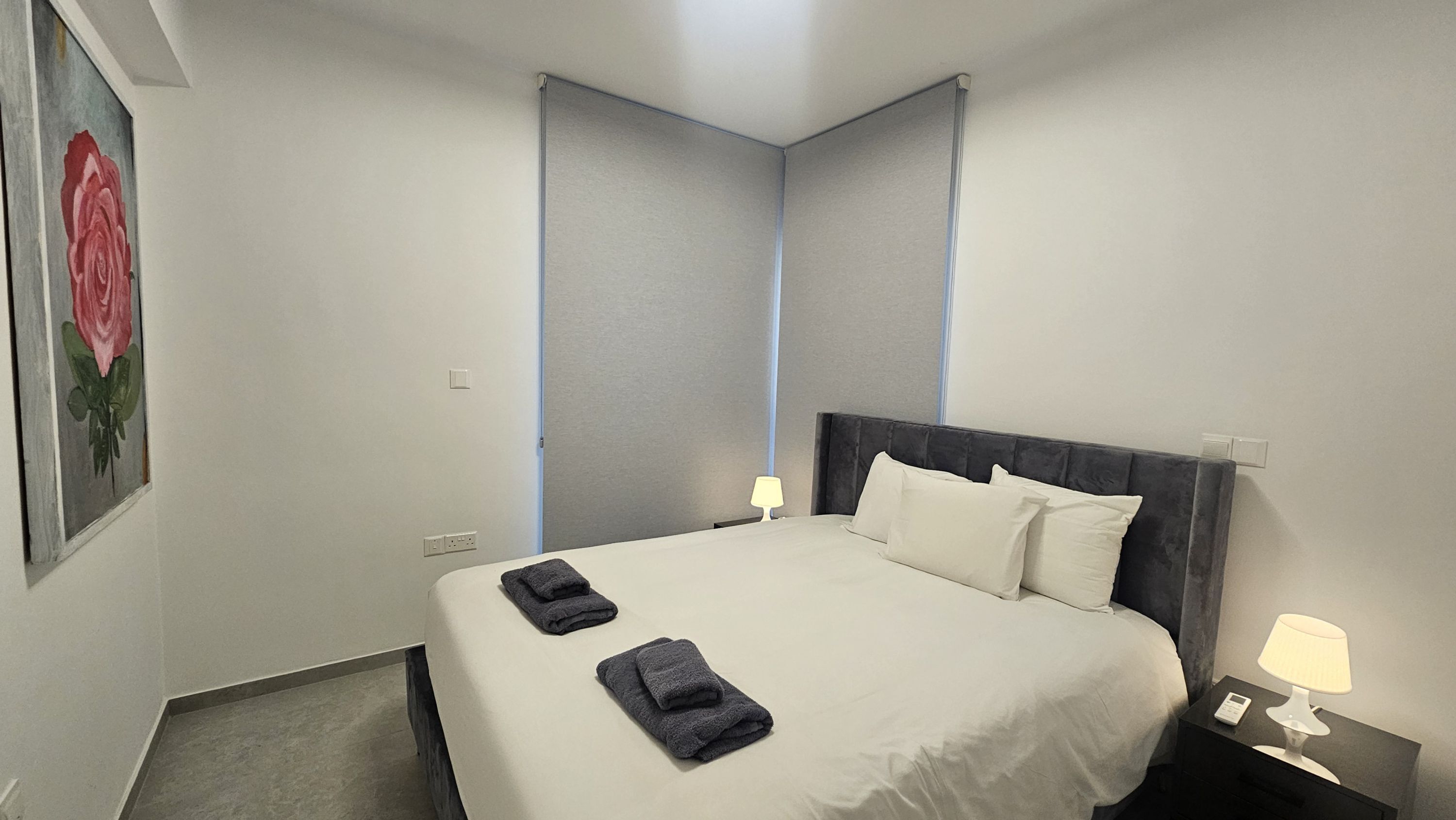 A hotel room with a bed, two bedside lamps, a nightstand, and a painting of a rose on the wall.