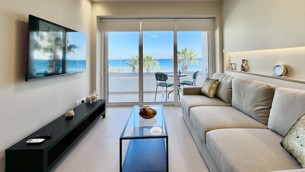 A modern living room with a sofa, a coffee table, a TV, and a balcony with a view of the sea. - 14 LCA - Stay Atalanta Apartment (Larnaca)