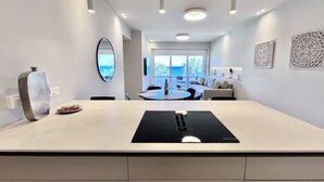 A modern kitchen with a central island, a dining area with a view, and a living space with a sofa and decorative wall art. - 14 LCA - Stay Atalanta Apartment (Larnaca)