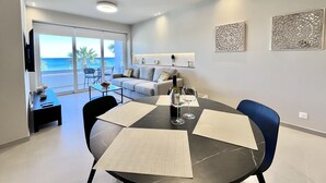 A modern living room with a dining table, sofa, and a view of the ocean through large sliding doors. - 14 LCA - Stay Atalanta Apartment (Larnaca)