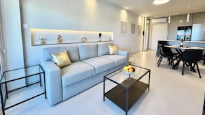 A modern living room with a grey sofa, a glass coffee table, and a dining area with a white table and black chairs. - 14 LCA - Stay Atalanta Apartment (Larnaca)