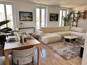 Ein modernes Wohnzimmer mit einem beigefarbenen Sofa, einem Esstisch für vier Personen und einem großen Fenster mit Blick auf die Stadt.