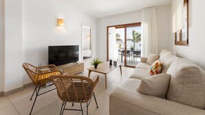 Un salon moderne comprenant un canapé, deux fauteuils en osier, un téléviseur, une petite table avec une plante et une porte-fenêtre coulissante donnant sur un balcon.