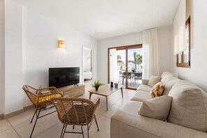 Una sala moderna con un sofá, dos sillones de mimbre, un televisor, una mesita con una planta y un ventanal corredizo que da a un balcón.