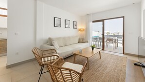 A modern living room with a sofa, two wicker chairs, a coffee table, and a TV. There is a balcony with outdoor furniture visible through the glass doors. - Villa Los Altos Yaiza 9A by Escape Home (Playa Blanca)