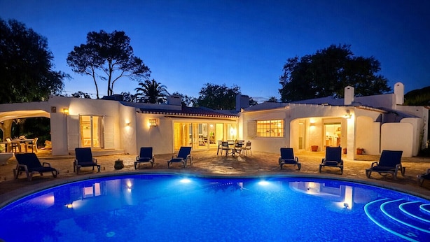 Une piscine entourée de chaises longues et d’un espace patio, le tout bordé d’un bâtiment dont les portes sont ouvertes et où les lumières sont allumées.