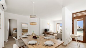 Ein moderner Essbereich mit einem Holztisch, gedeckt für vier Personen, weißen Stühlen, einer Hängelampe und Blick ins Freie durch eine Tür.