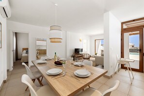 A modern dining area with a wooden table set for four, white chairs, a hanging light fixture, and a view of the outdoors through a door. - Villa Los Altos Gadea 9B by Escape Home (Playa Blanca)