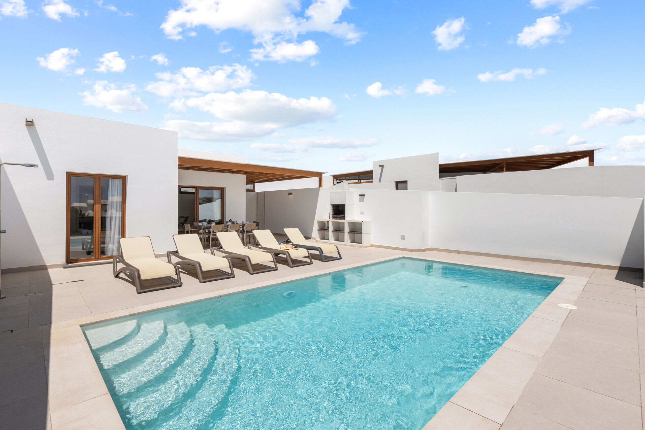 A rectangular pool with lounge chairs, a wooden pergola, and a modern white building with a glass door.