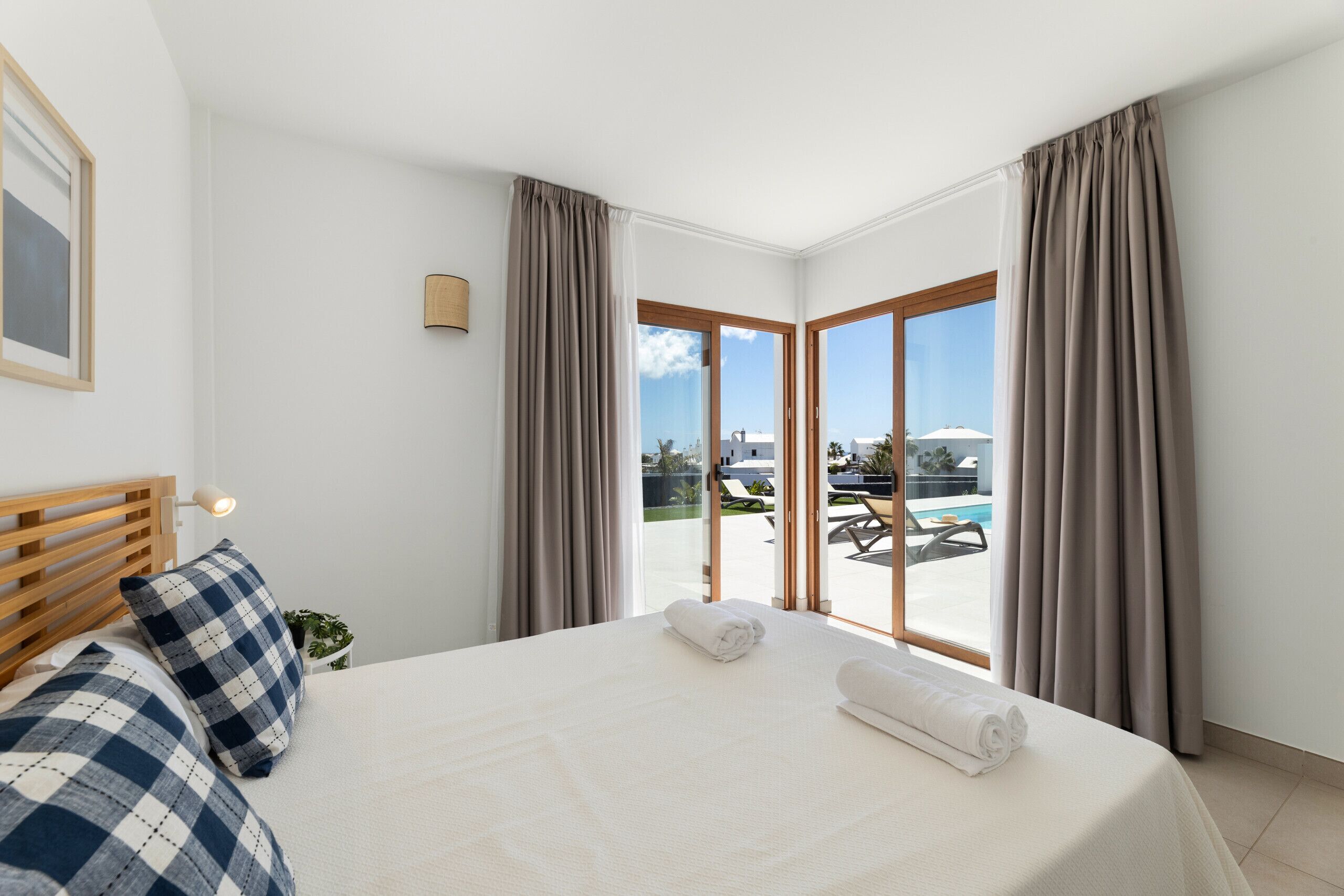 Habitación de hotel con cama, cabecera, lámpara de noche, ventana con cortinas y vistas a una zona de piscina exterior.