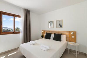 A hotel room with a bed, a small table, a window with curtains, and two framed pictures on the wall. - Villa Los Altos Navya 13B by Escape Home (Playa Blanca)