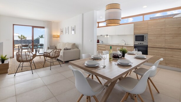 Et moderne køkken med træskabe, et spiseområde med et træbord og hvide stole, samt en balkon med udsigt til palmer.