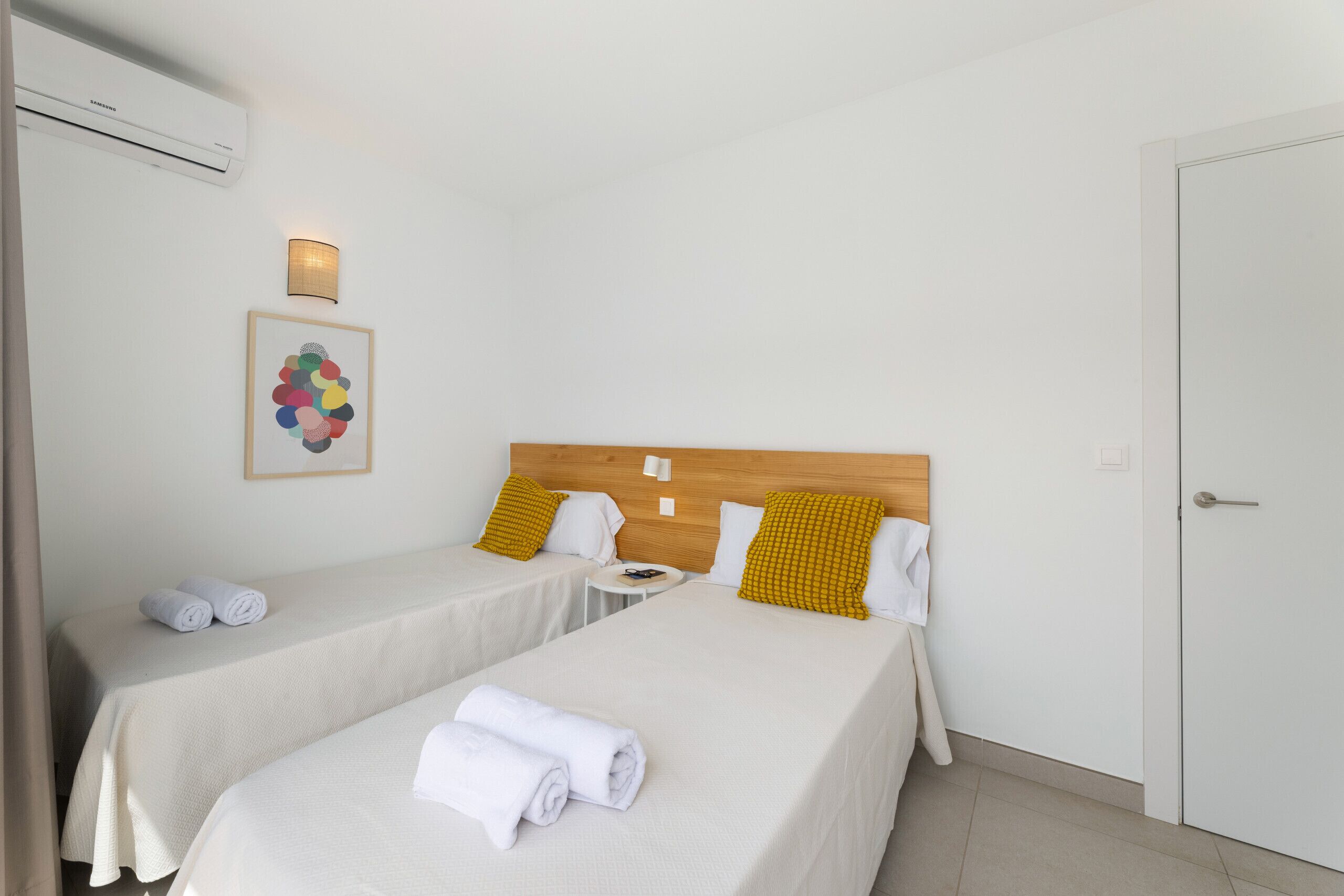 A hotel room with two beds, a wooden headboard, a wall-mounted air conditioner, a small table, and a framed picture.