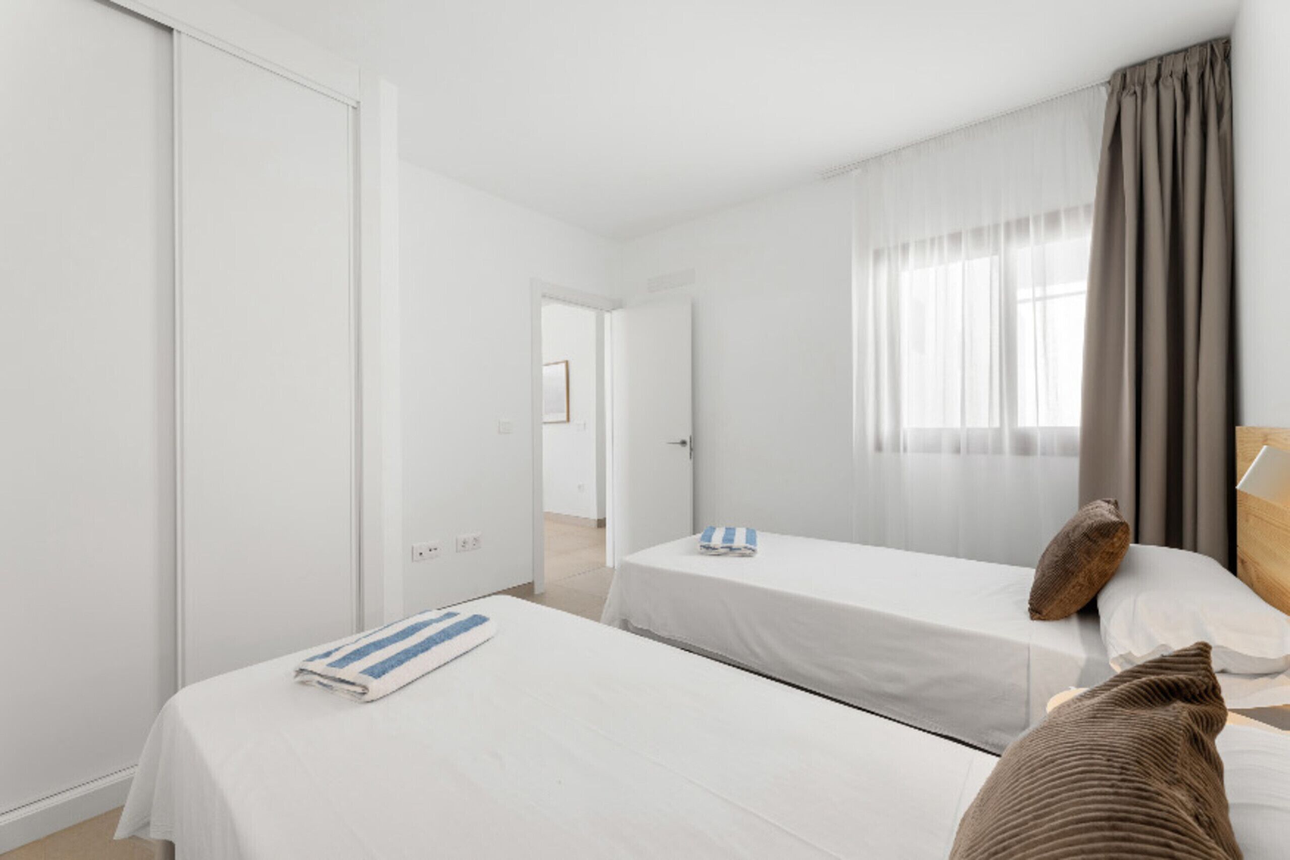 A hotel room with two single beds, a large window with curtains, and a door leading to another room.