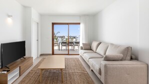 A modern living room with a flat-screen TV, a wooden coffee table, a beige sofa, and a large sliding glass door leading to an outdoor patio with a table and chairs. - Villa Los Altos Deva 12A by Escape Home (Playa Blanca)