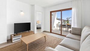A modern living room with a flat-screen TV, a wooden coffee table, and a sofa. There is a sliding glass door leading to an outdoor patio with a table and chairs. - Villa Los Altos Deva 12A by Escape Home (Playa Blanca)