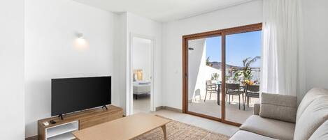 A modern living room with a flat-screen TV, a wooden coffee table, and a sofa. There is a sliding glass door leading to an outdoor patio with a table and chairs.