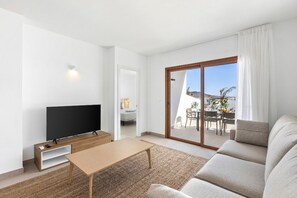 A modern living room with a flat-screen TV, a wooden coffee table, and a sofa. There is a sliding glass door leading to an outdoor patio with a table and chairs. - Villa Los Altos Deva 12A by Escape Home (Playa Blanca)