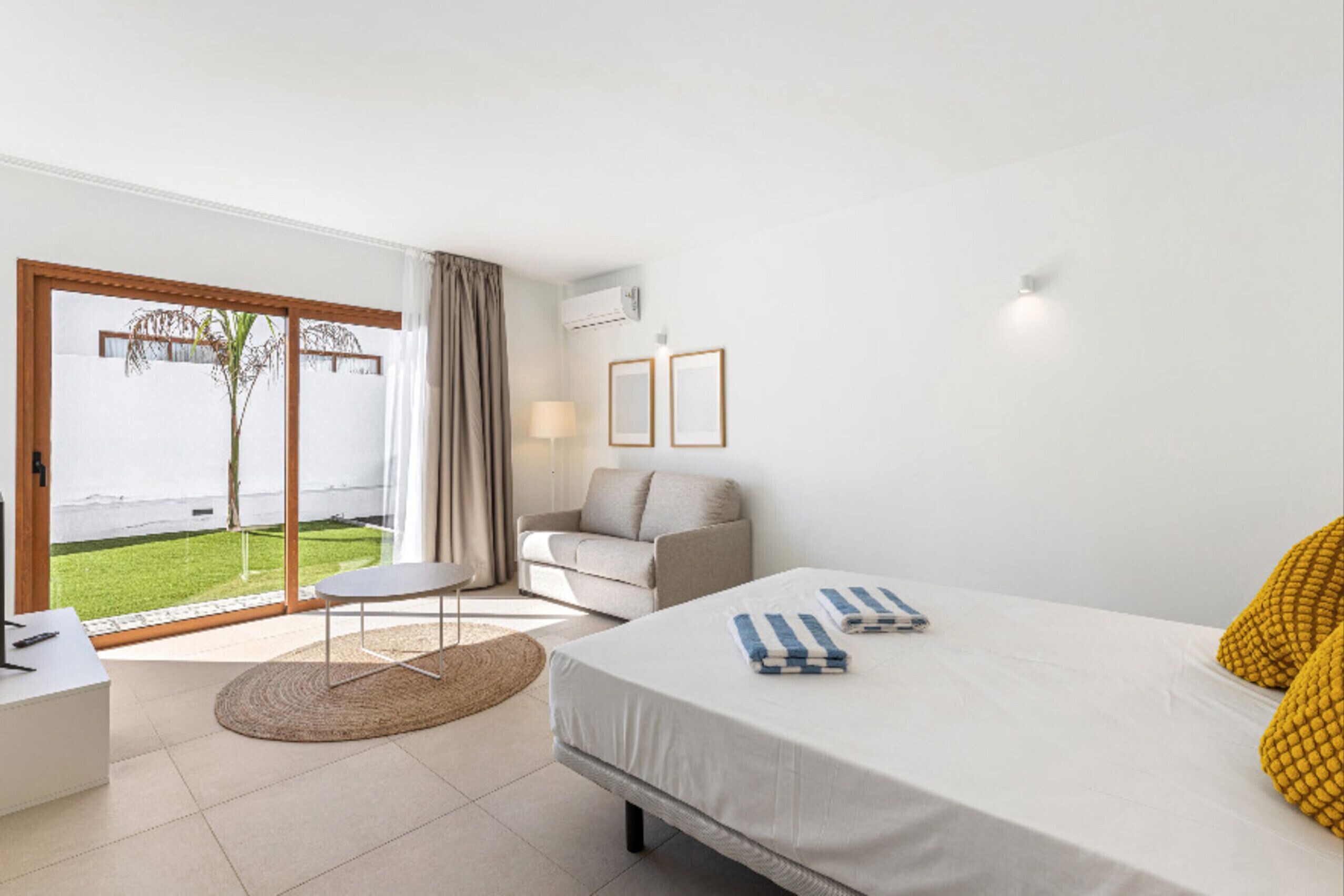 A modern hotel room with a bed, a sofa, a small table, and a view of a garden through a sliding door.