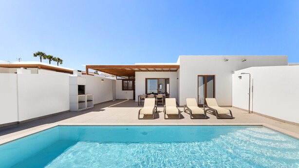 A modern pool area with lounge chairs, a clear pool, and a building with large windows and a wooden ceiling. - Villa Los Altos Blanca 8A by Escape Home (Playa Blanca)