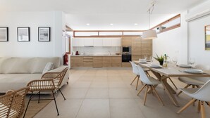 A modern kitchen with wooden cabinets, a dining area with white chairs, and a living room with a sofa and wicker chairs. - Villa Los Altos Blanca 8A by Escape Home (Playa Blanca)