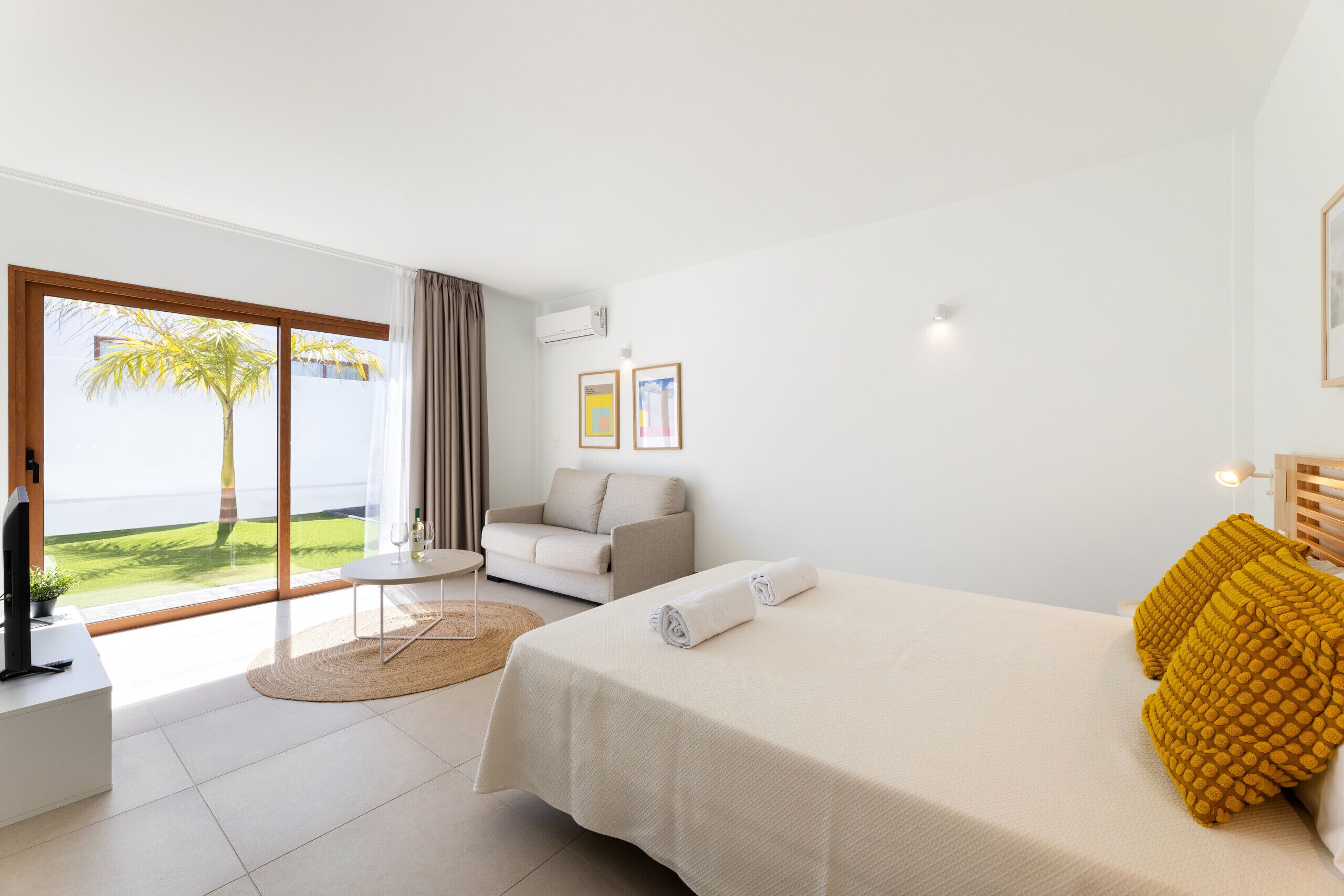 Una habitación de hotel moderna con una cama, un sofá, una mesita, un televisor y un ventanal con vistas a palmeras.