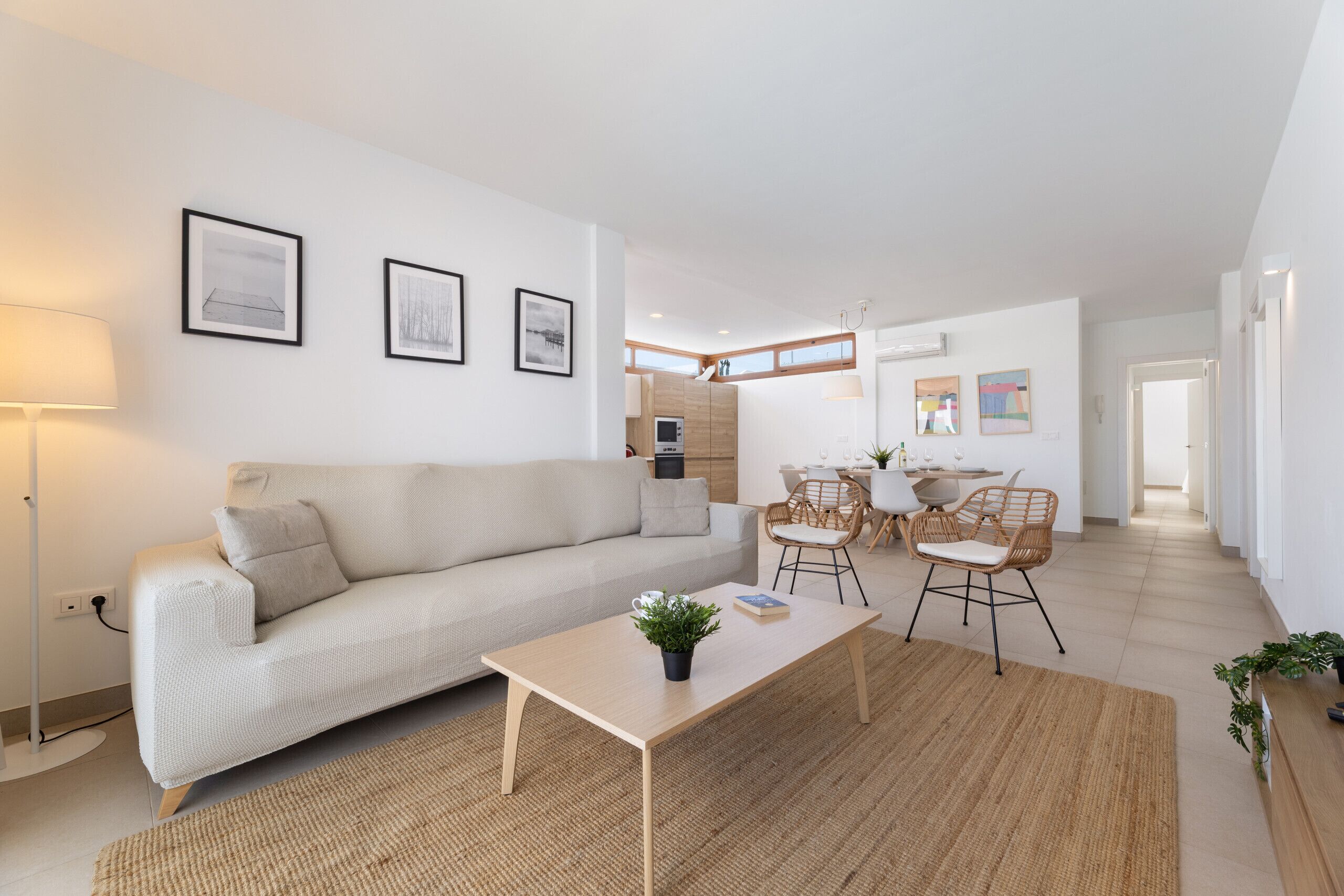 Una sala moderna con sofá, mesa de centro y sillones, un comedor con mesa y sillas, y una cocina al fondo.