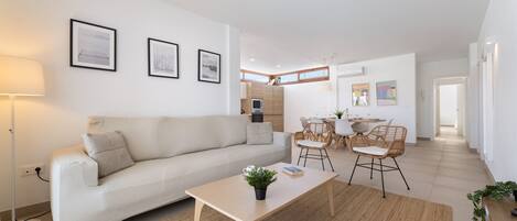 Una sala moderna con sofá, mesa de centro y sillones, un comedor con mesa y sillas, y una cocina al fondo.