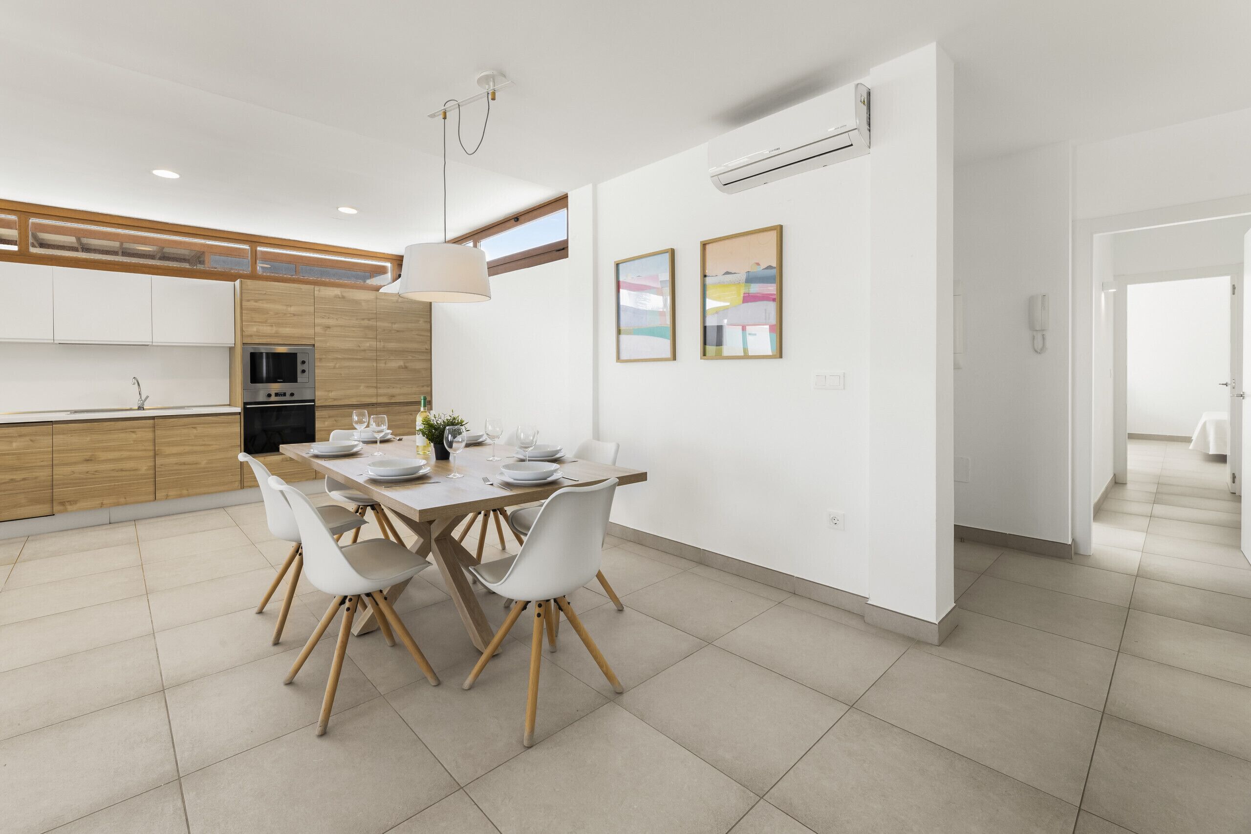 A modern kitchen with white cabinets, a dining table with chairs, and an air conditioning unit on the wall.