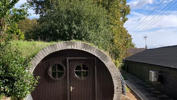 A circular, dark wooden structure with two windows, surrounded by greenery and a paved walkway. - Nice Holiday Home in Hastingleigh (Ashford)