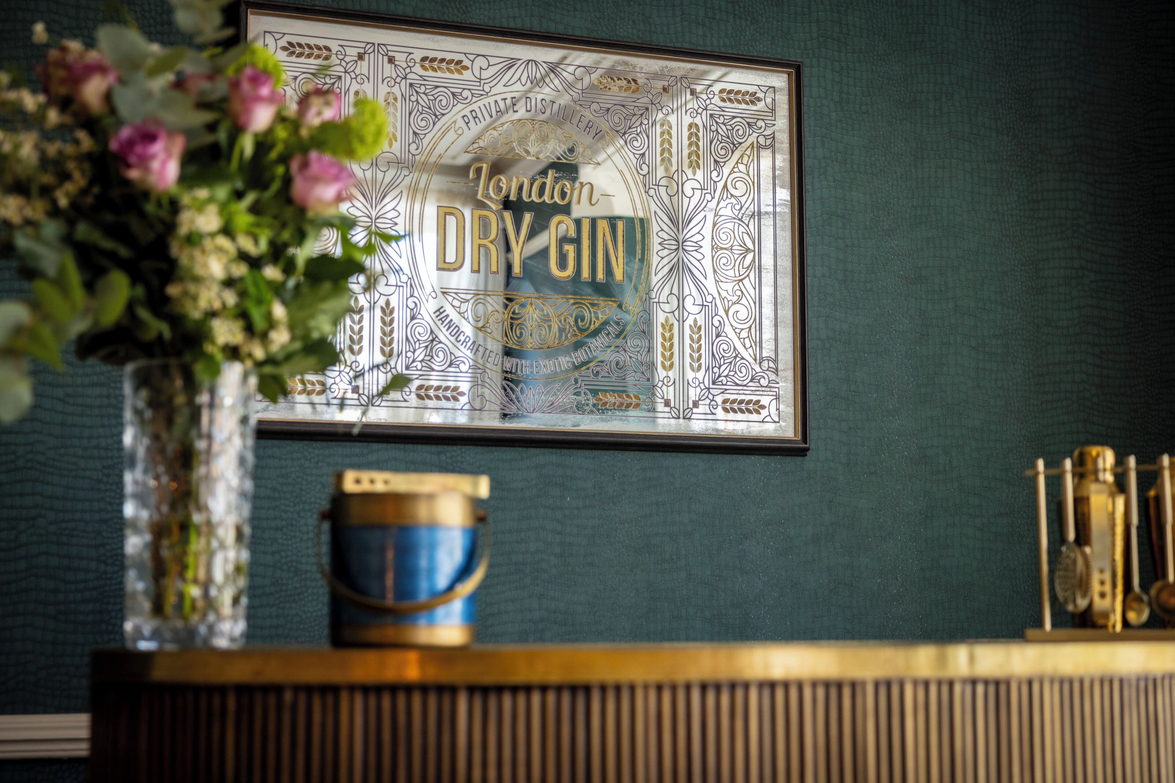 A framed sign with 'London Dry Gin' text, a vase with flowers, a blue container, and a gold-colored object on a counter.