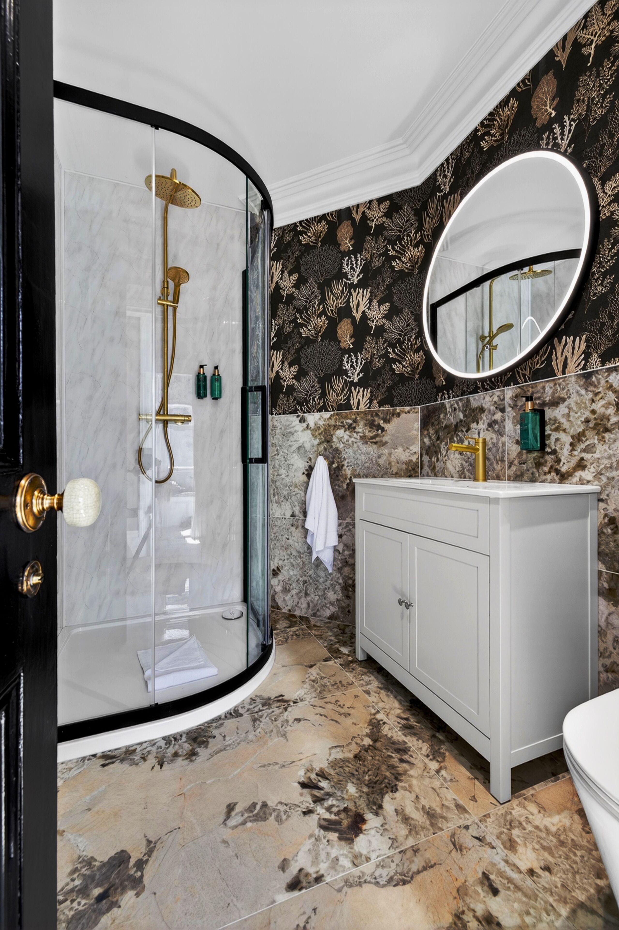 A bathroom with a glass-enclosed shower, a white vanity, a round mirror, and a patterned wall.