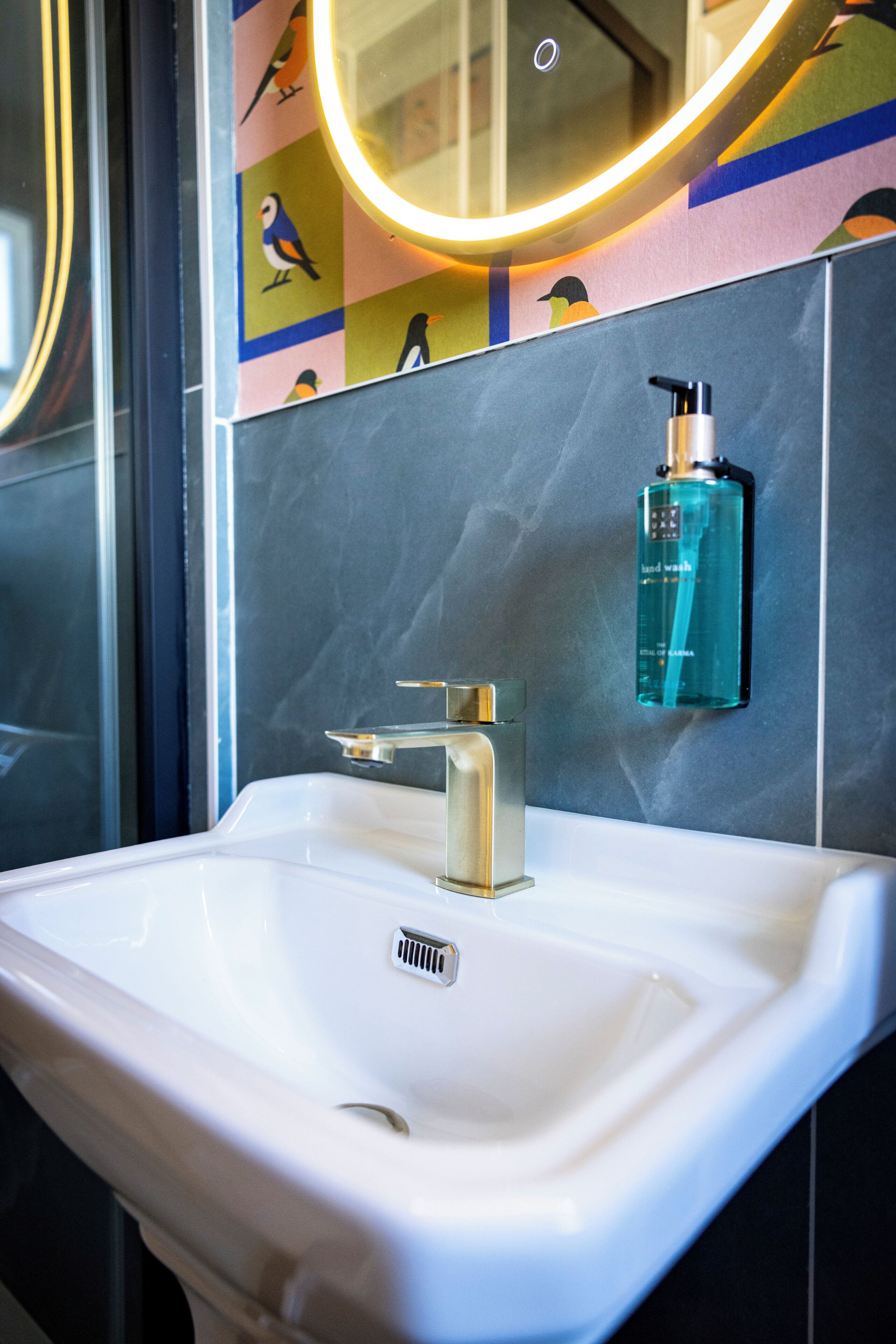 A bathroom with a white sink, a modern faucet, a handheld soap dispenser, and a wall-mounted mirror with a circular light.