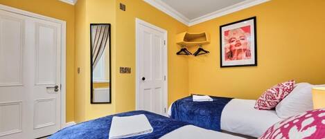 Habitación de hotel con dos camas, paredes amarillas, puertas blancas, un cuadro enmarcado y una toalla sobre una de las camas.