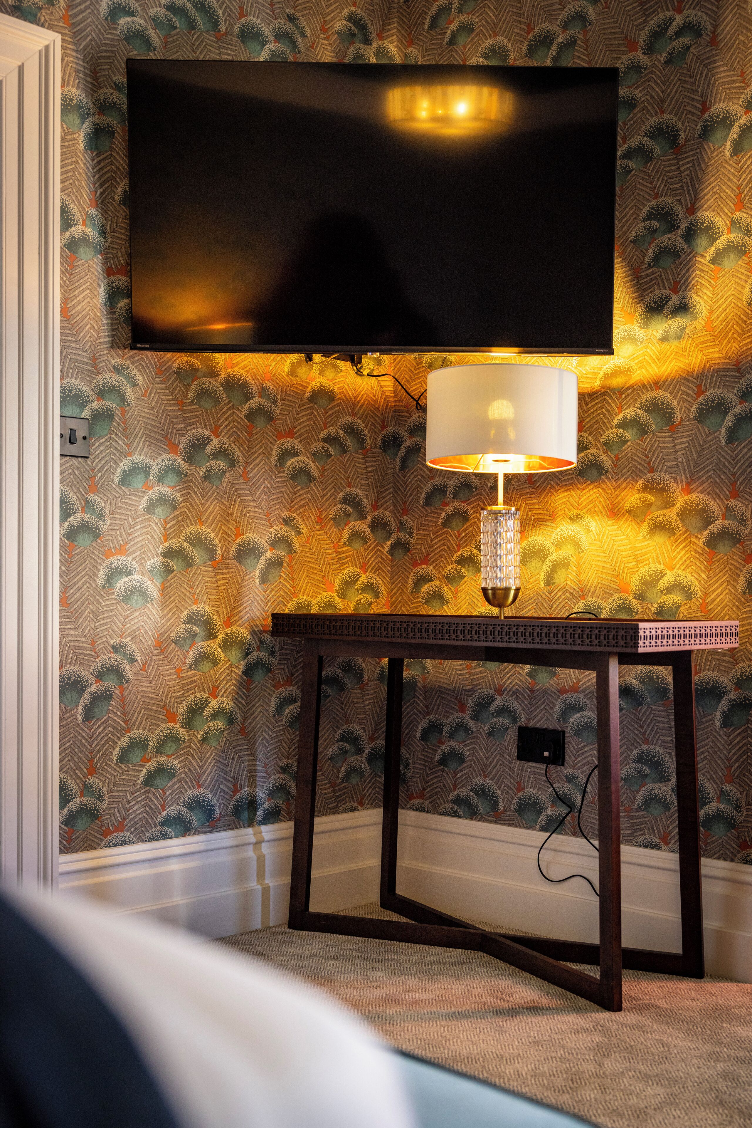 A television mounted on a wall with a patterned wallpaper, a table with a lamp, and a carpeted floor.