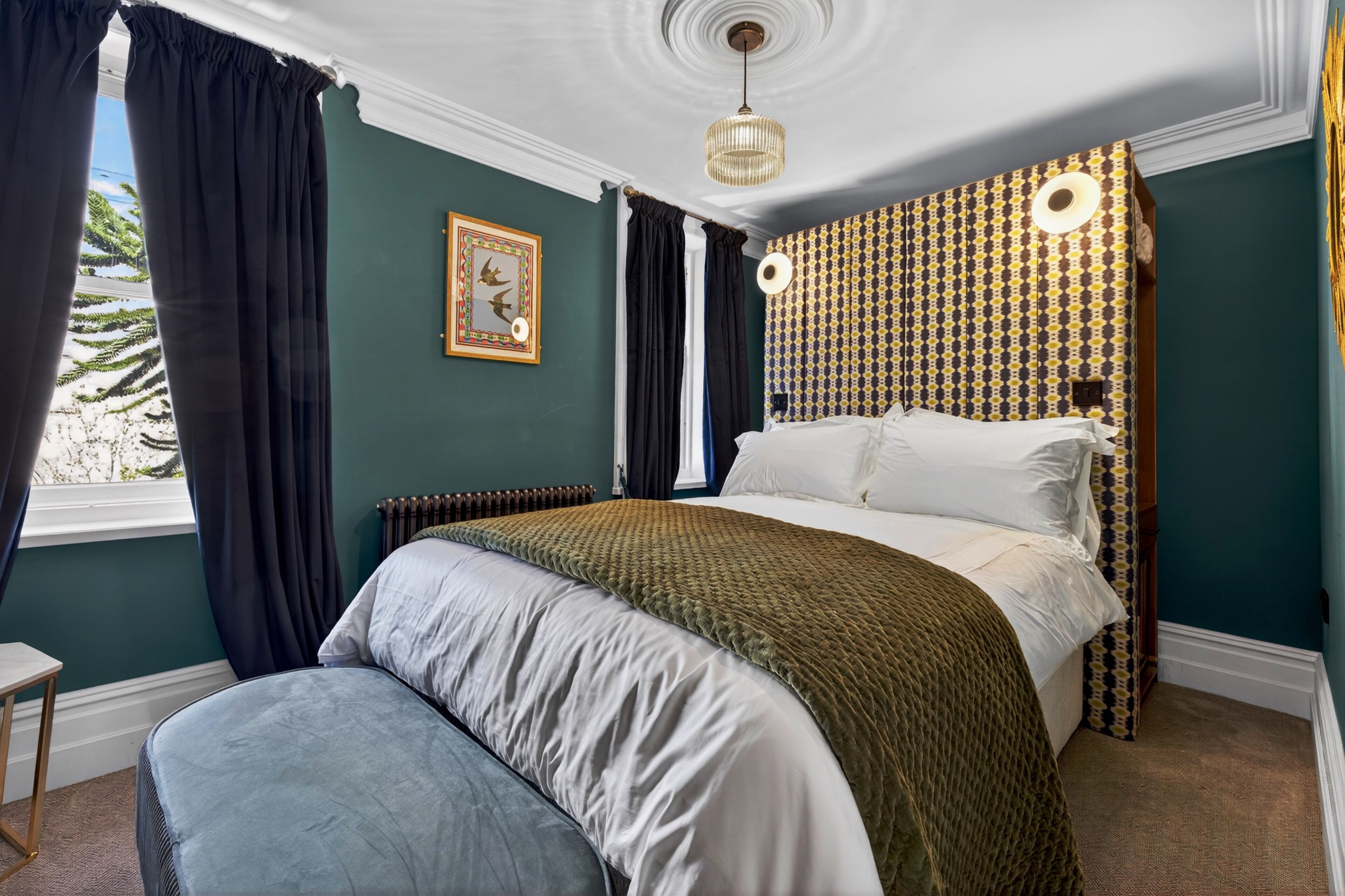Un dormitorio con cama, mesita de noche, radiador, ventana con cortinas y pared con diseño.