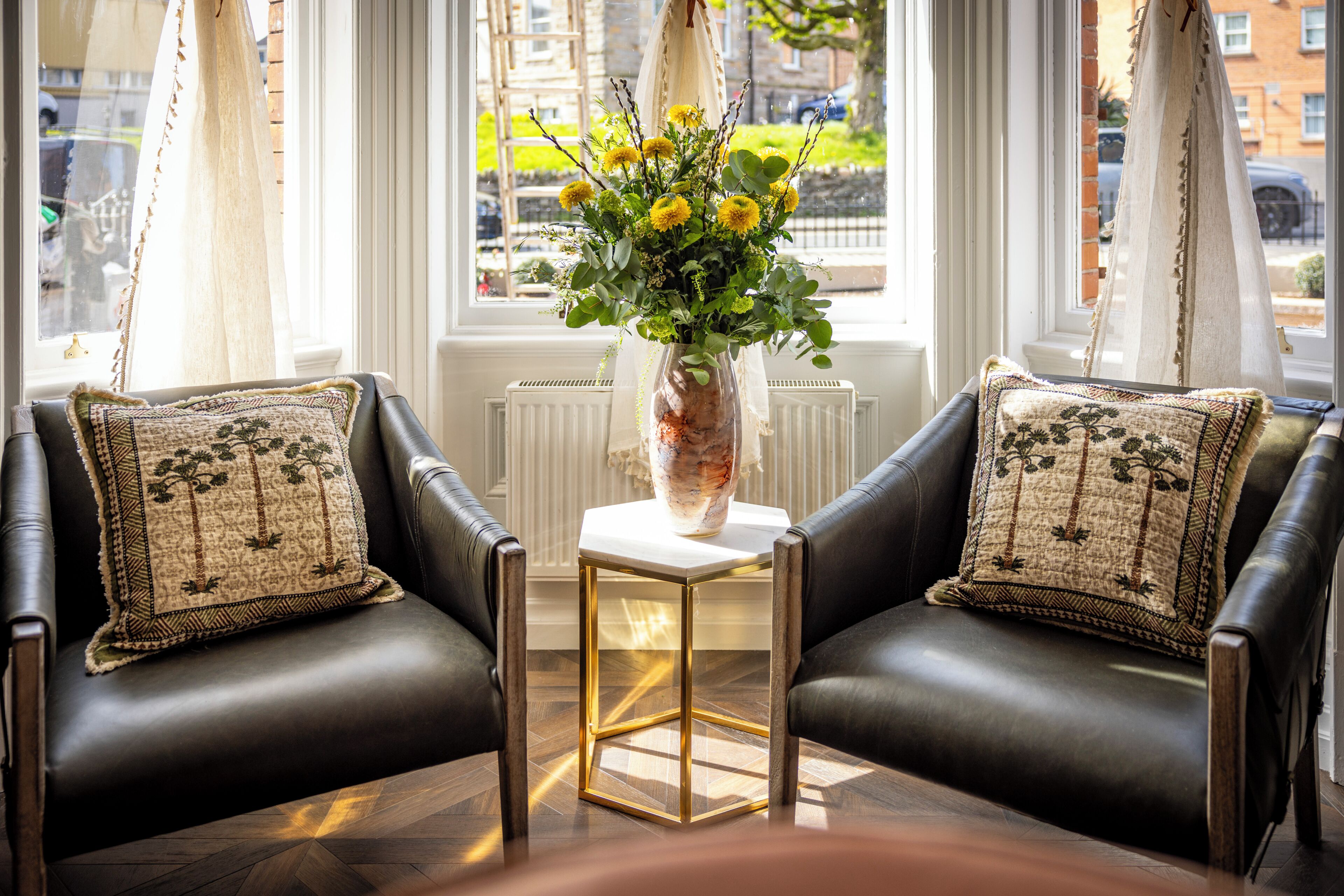 A room with two leather armchairs, a white vase with yellow flowers, and a view of the street through large windows.