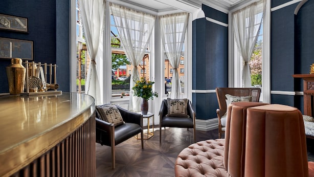 A room with a bar, two leather chairs, a tufted ottoman, and large windows with curtains. - Clarence House (Londonderry)