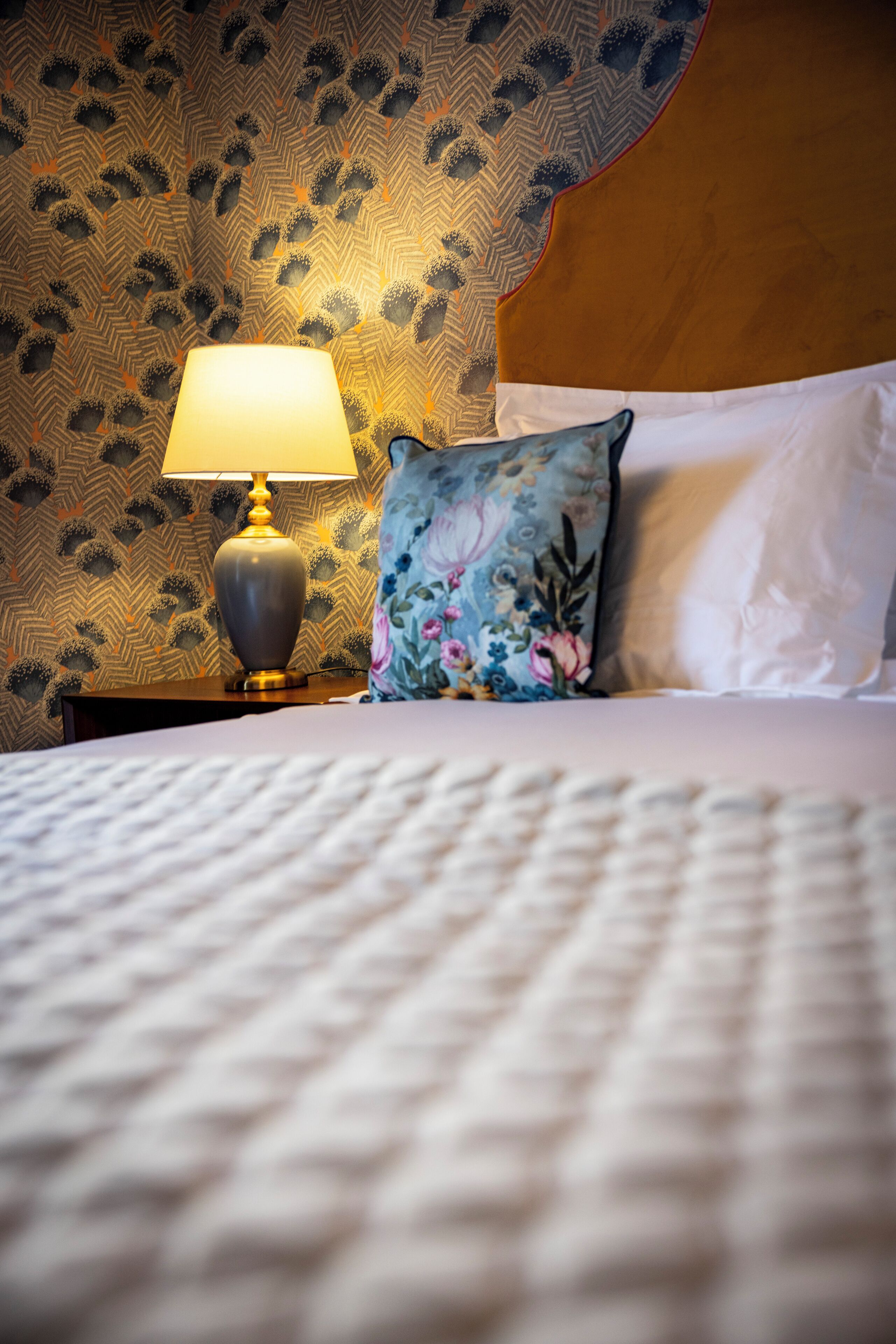 A neatly made bed with a patterned wall and a nightstand with a lamp.