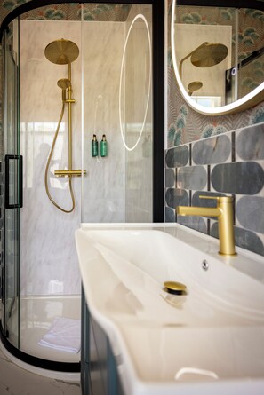 A modern bathroom with a glass-enclosed shower, a white sink with gold fixtures, and a mirror with a gold frame. - Clarence House (Londonderry)