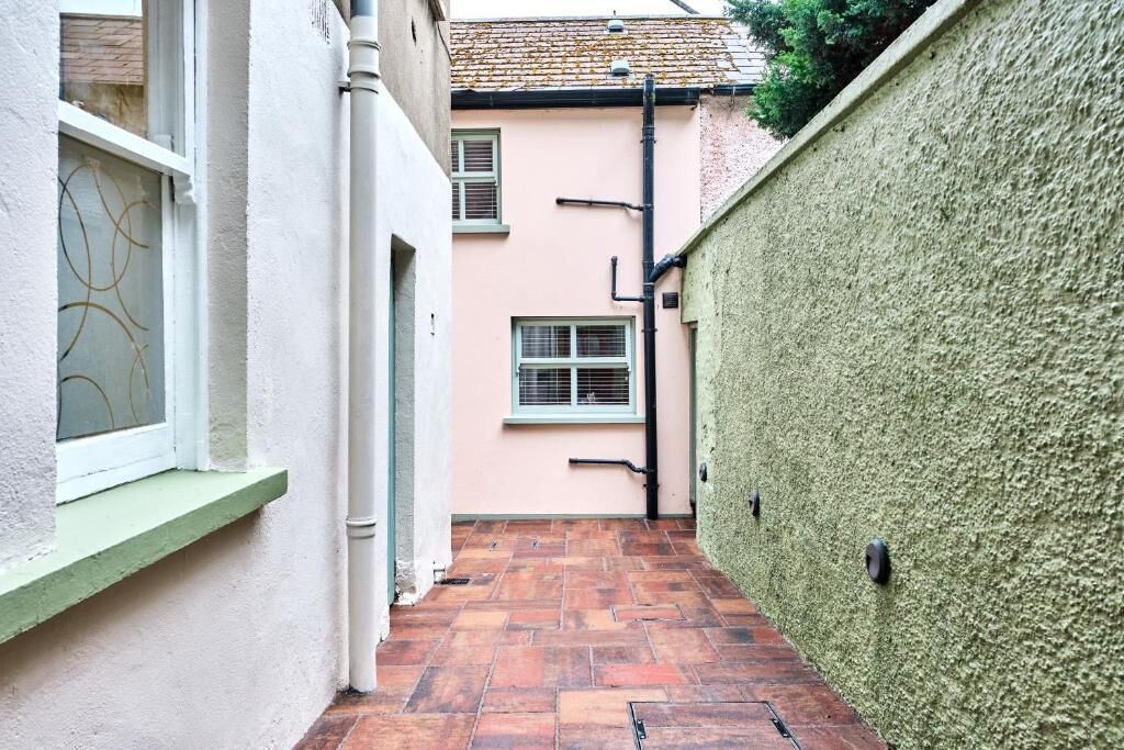 A narrow alleyway with a tiled floor, a green wall on one side, and a building with a pink facade and windows.