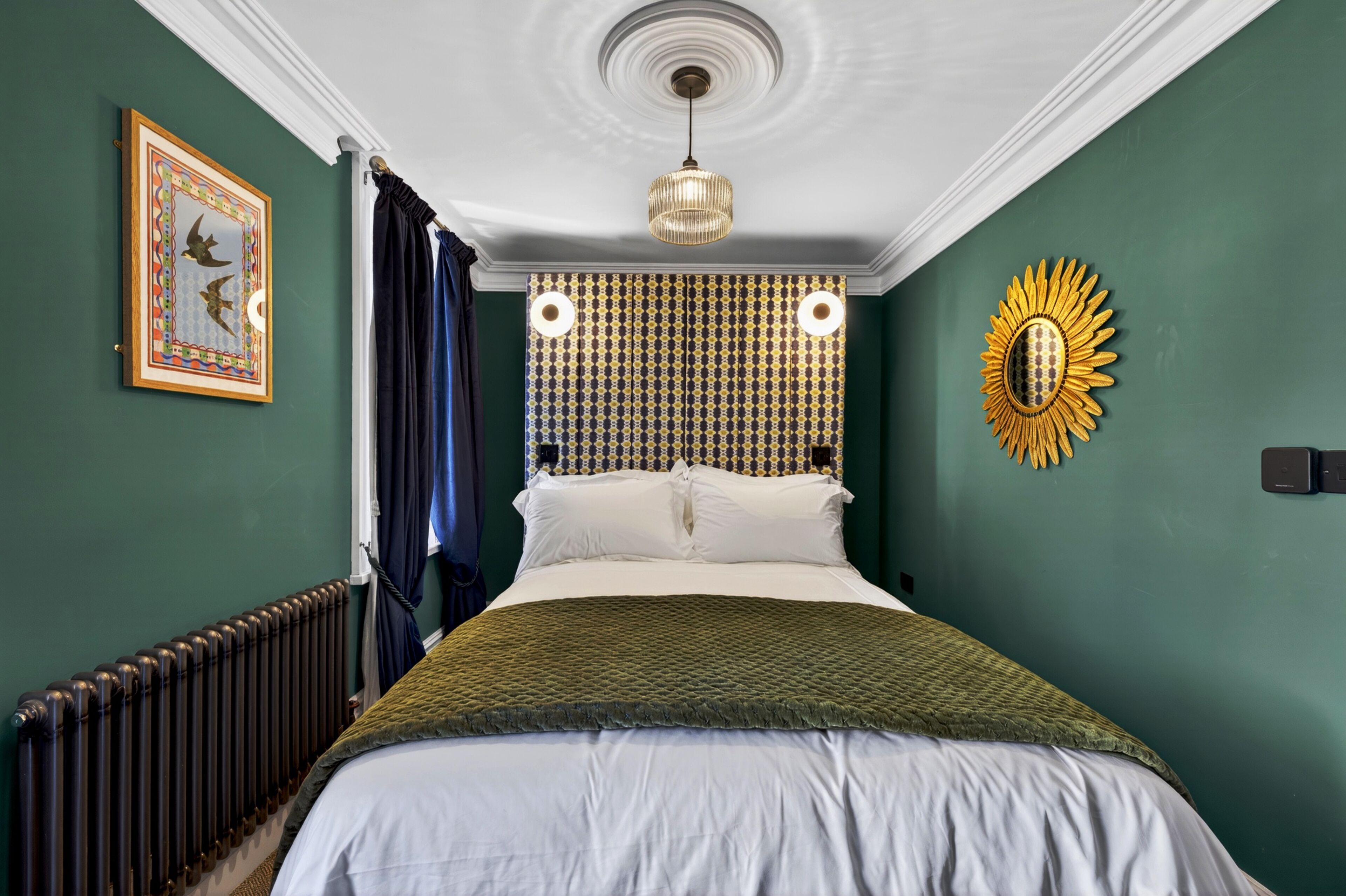 A bedroom with a bed, a radiator, a ceiling light, a wall clock, and a sunflower decoration.