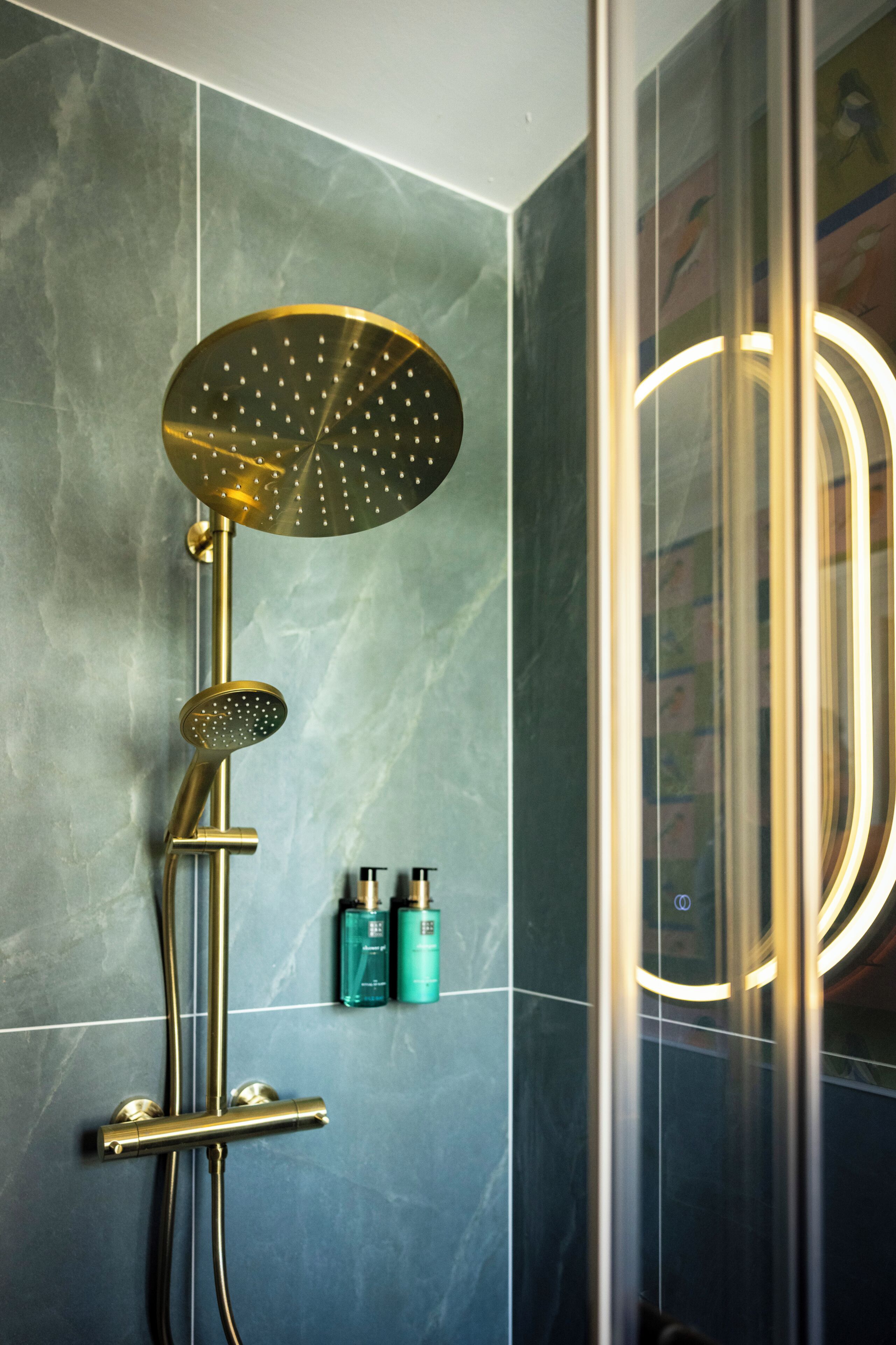 A modern bathroom with a gold shower head, a handheld shower, and a glass-enclosed mirror with lights.