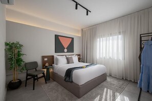 A modern hotel room with a bed, a chair, a nightstand, a potted plant, and a painting on the wall. - Livingville East (New Cairo)