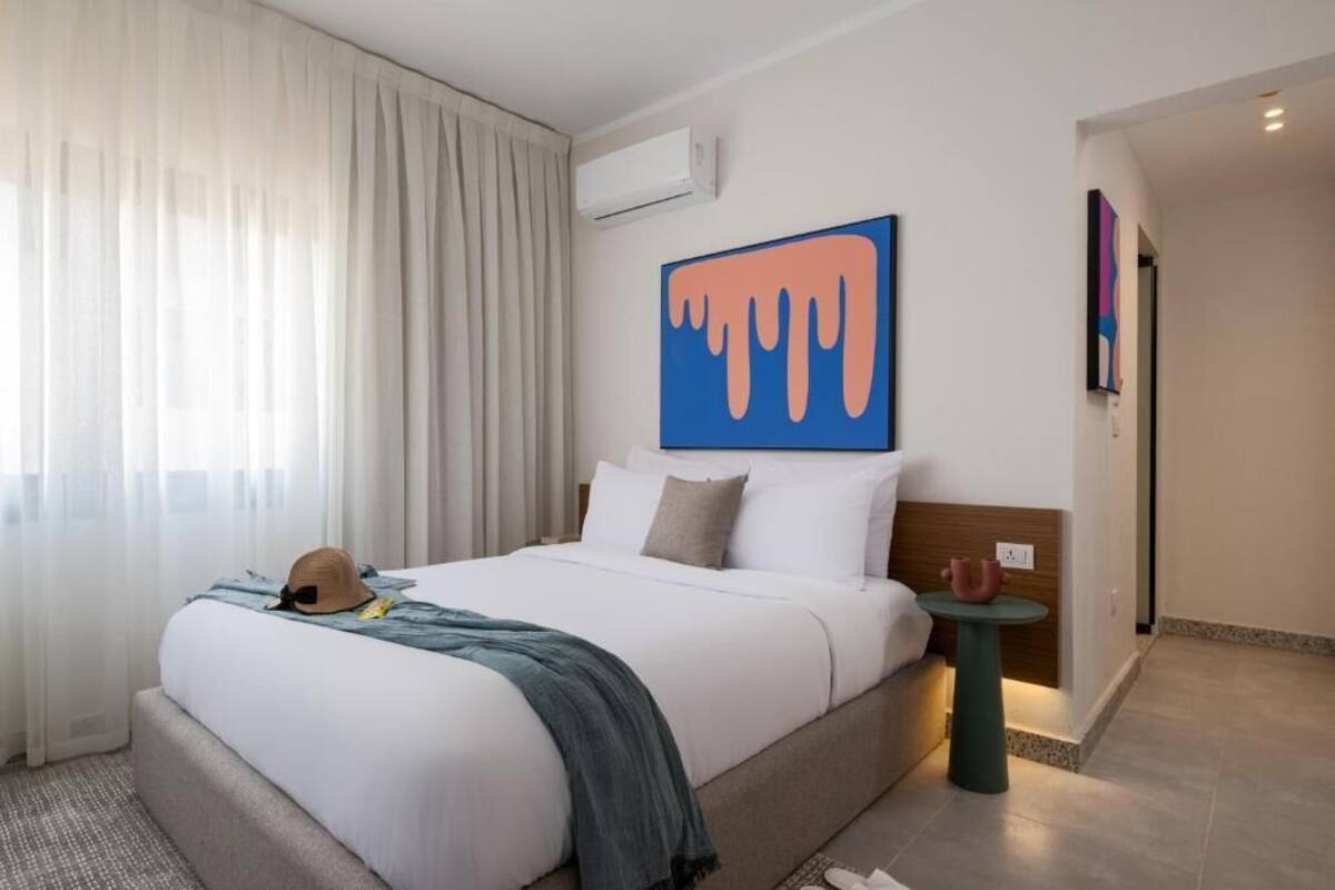 A hotel room with a large bed, a small side table, a wall-mounted air conditioner, and a painting above the bed.