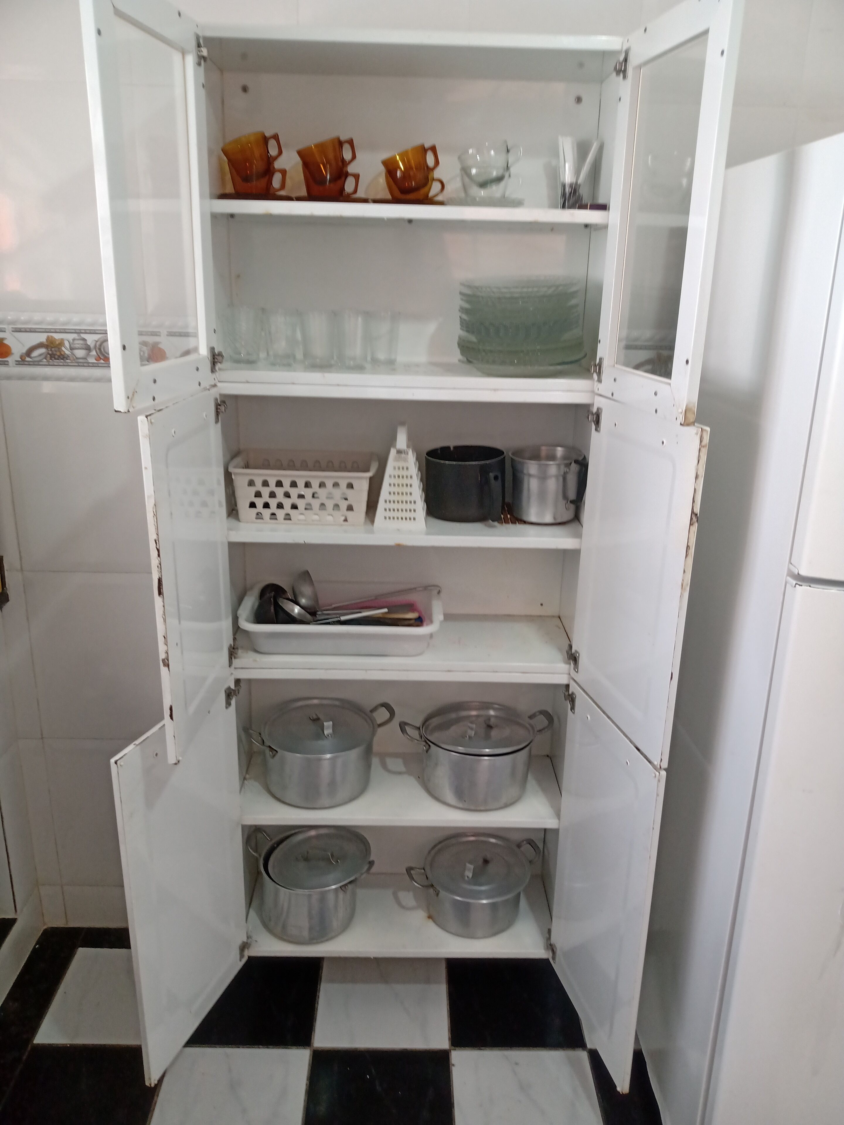 Eine Küche mit einem weißen Kühlschrank, einem Schachbrettboden und Regalen mit Töpfen, Pfannen und Küchenutensilien.